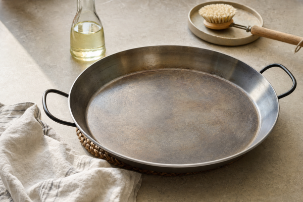 La Ideal paella pan on a kitchen worktop beside neutral oil, a cotton cloth and a cleaning brush.